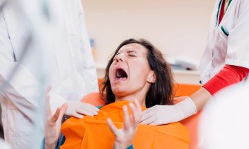תמונה של המאמר רשלנות רפואית בטיפולי שיניים בה רואים אישה צועקת בכיסא רופא שיניים פיה פתוח לרווחה שני רופאי שיניים עם כפפות בודקים את פיה תאורה בהירה במרפאת שיניים