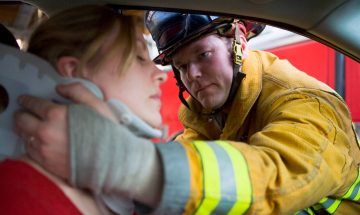 תמונת נושא של המאמר עורך דין תאונת דרכים עם נפגעים המציג כבאי בחליפה צהובה מניח צווארון אורתופדי לצווארה של אישה במושב הנהג לאחר תאונת דרכים המייצג חילוץ וטיפול ראשוני