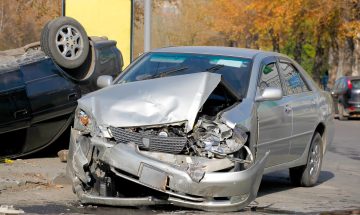 תמונת נושא של המאמר על עורך דין תאונות דרכים בנתניה המציגה תאונת דרכים קשה שבה מכונית כסופה עם חזית מעוכה נמצאת לצד מכונית כהה שהתהפכה על צידה התאונה התרחשה בכביש מרופש עם עצים בשלכת ברקע.