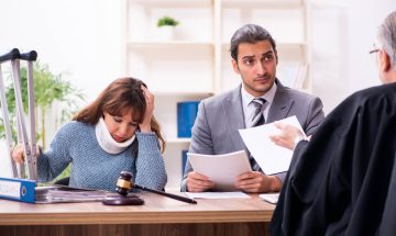 תמונת נושא של המאמר על עורך דין לנפגעי עבודה המציגה אישה עם צווארון רפואי וכתף תפוסה יושבת ליד שולחן עם עורך דין שפניו אל שופט או עורך דין בכיר נוסף השולחן עמוס בתיקים ופטיש שופטים יש קביים ברקע.